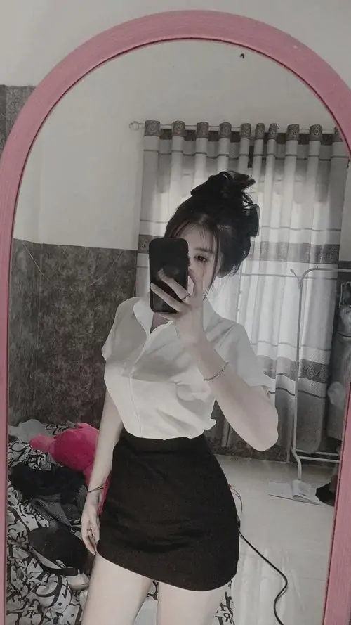 Tấm gương dọc phản chiếu cô gái mặc áo trắng và chân váy đen, dáng nghiêng gọn gàng trong không gian phòng ngủ