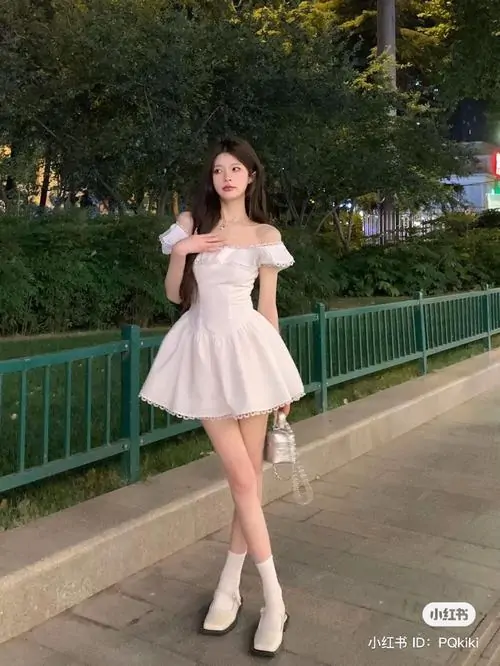Góc chụp toàn thân ghi lại hình thiếu nữ diện váy ngắn trắng xòe, tóc dài buông mềm, tạo dáng nhẹ nhàng bên lan can xanh