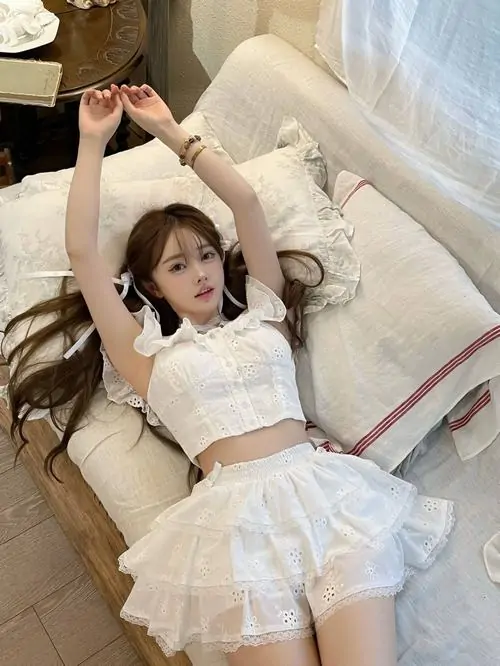 Trên giường phủ ga trắng, gái xinh mặc váy ngắn màu kem nằm thư giãn, mái tóc nâu xõa nhẹ và nụ cười thoải mái