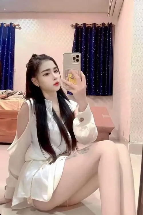 Trên giường ngủ phủ ga hồng, cô gái mặc váy trắng ngắn ngồi chống tay, mái tóc đen dài và biểu cảm nhẹ nhàng