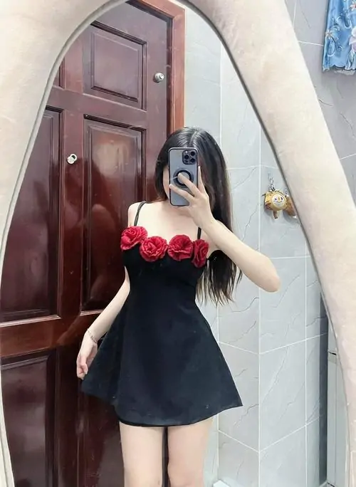 Trước cửa gỗ, cô gái mặc đầm đen ngắn điểm hoa đỏ, dáng đứng nghiêng cùng điện thoại tạo cảm giác đời thường