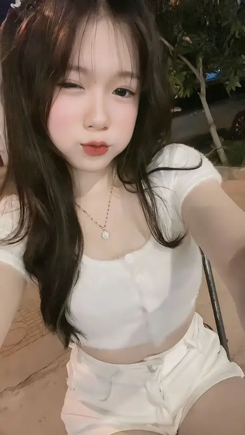 Cô gái mặc áo trắng ngồi trong bóng cây, tóc đen thả dài, ánh mắt nhìn máy nhẹ nhàng và nền tối làm mặt sáng hơn