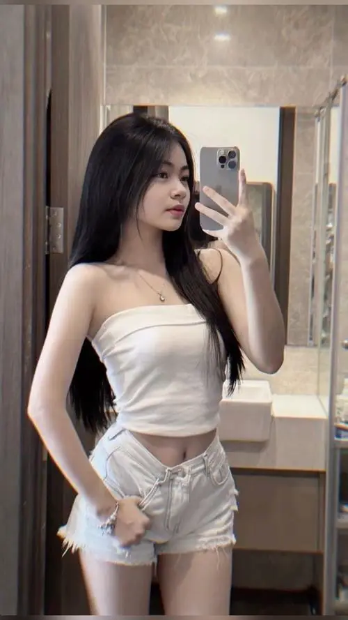 Trước gương trong phòng thay đồ, cô gái mặc váy trắng ôm dáng, tóc dài thẳng và tư thế đặt tay đầy tự tin